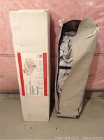 Box standing next to the beige storage bag containing the bed parts in a basement environment.