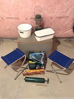 Wide view of camping lot showing cooler, lantern, enamel dishes, stools, air mattress box, and hand pumps.