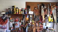Overview of pegboard with various hand tools including saws, wrenches, hammers, screwdrivers, and pliers hanging, plus some hardware packets and an orange extension cord coiled below.