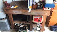 Wooden workbench with visible wear, 3 shelves beneath. Various items stored underneath. Mounted red Wilton Scout 3.5 jaw vise on front left corner.