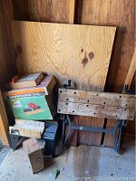 Photo showing the Black & Decker Workmate portable workbench folded, several boxes including miter box and wooden boards behind it.