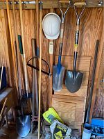 Wide shot of entire lot showing long handle tools hanging on wooden wall, safety helmet hanging, and bags on floor.