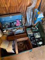 View of open green metal toolbox containing electrical outlets, switches, covers, and other parts plus cardboard box with coiled metallic materials.