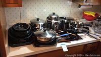 Overview of assorted cookware on countertop showing stack of black speckled roasting pans and stainless steel pots and pans with lids