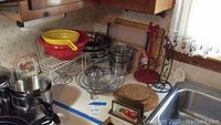 Top view of kitchen counter with various metal cooking items including colanders, wire baskets, cutting boards, decorative stands, and a yellow pan.