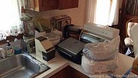 Overview of kitchen counter with various small kitchen appliances grouped together showing the lot contents including toaster oven, Rival ice crusher, Crock pot, waffle makers, and George Foreman grill.