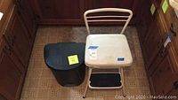 Black plastic trash can with lid and white step stool chair with two black steps next to it in a kitchen setting