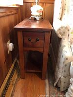 Wooden end table with drawer and open lower shelf placed beside a couch and wooden wall paneling.