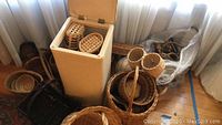 Photo showing cream-colored hinged lid wicker laundry hamper filled with a couple of smaller wicker baskets, with several wicker baskets arranged around it on the floor near a curtain.