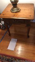View of one wooden side table showing top surface, single drawer with metal handle, and Queen Anne feet on hardwood floor with lamp on top.