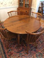 Round wooden dining table shown with chairs around it on Persian-style rug inside a room with floral wallpaper.
