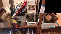 Wide view showing several plastic bins with assorted picture frames standing upright and lying down, various gift bags stored and plastic storage containers full of items.