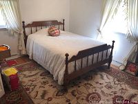 Full view of the bed frame made from wood with turned posts and spindle design on headboard and footboard, with white coverlet and round plaid throw pillow on top.