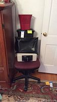 Maroon office chair with fireproof lockbox, black bag on top, white base, and red trashcan stacked on top of chair in corner of room.