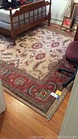 Full view of the woven area rug placed on wood floor under bed and chair, showing beige background with large floral design and red border