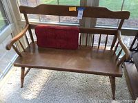Wooden bench with slatted backrest, natural wood finish and rounded armrests seen next to window with a red rectangular tufted pillow on seat.