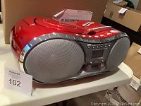 Side angled view of Emerson red portable boombox showing speaker grill, CD player, control buttons, and carrying handle on top. Unit is sitting on a table.