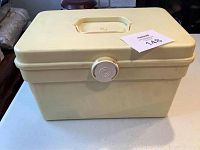 Closed view of the cream plastic sewing box with handle and circular latch.