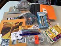 Photo showing two contemporary clipboards, student clock, academic writing manual, exercise books, pens, and a glue gun.