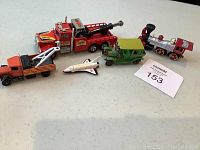 Five toy vehicles displayed on a white surface, including a red and orange tow truck, white space shuttle, green vintage car with yellow roof, red tractor-train car, and a red fire truck.