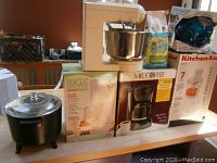 Wide view of all kitchen appliances on table including KitchenAid mixer, Magic Bullet box, Mr. Coffee box, KitchenAid food processor, black cooker with glass lid
