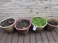 Four outdoor planters in a row against brick wall, filled with soil and one with green plants.