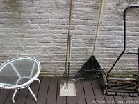 Photo showing glass topped circular side table and gardening tools including a metal dustpan, leaf rake, and pitchfork.