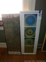 Two framed prints standing upright near a window, one with four geometric blue and green spirals in a white frame and the other a Gustav Klimt landscape print with floral foreground.