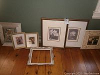 Seven framed floral artworks and one empty ornate frame displayed on wood floor against green wall.