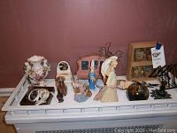 Photo of assortment of various decorative items arranged on a white table against a brown wall, showing floral vase, figurines, glass ball, and other decor pieces.