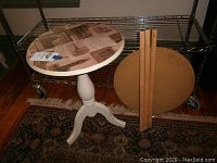 Photo showing white round bedside table in front, and round wooden table top with detached legs behind on metal rack.