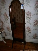 Full front image of a Bombay standing mirror with wooden frame and angled legs in a floral wallpapered room.