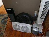 Multiple fans displayed on wooden floor near a wall outlet including white tower fan, black floor fan, box fan, and small heater.