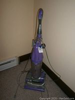 Full view of purple Dyson Animal upright vacuum cleaner standing on carpet near wall, power cord attached.
