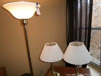 Three brass lamps together: one tall floor lamp and two smaller swing arm table lamps with white pleated shades