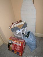 Photo showing ironing board leaning against wall, plastic storage bin, assorted hangers scattered on top, carpet step treads folded on storage bin, fabric bag on floor.