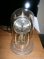 Front and slight angled view of Bulova anniversary clock under glass dome, showing gold-tone face with Roman numerals, ornate detailing, and crystal base.