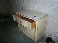 Front view of beige wooden dresser showing single drawer and double-door cabinet below, with ornate handles and curved design.