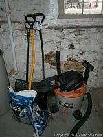 Wide shot showing RIDGID wet dry vacuum, two snow shovels, and bags of ice melt salt on the floor in a basement corner.