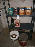 Plastic shelving unit with multiple cans and containers of wood sealers, waterproofing, deck stain, mold remover, and two large buckets on bottom shelf.