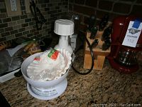 Photo showing a white kitchen scale with a coffee filter holder on top placed on a granite kitchen counter near a wooden knife block with knives.