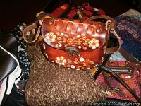 Brown leather purse with floral embroidery on a woven top flap, resting on knitted items and other bags