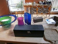 Full view of collection on a table including the wine bottle holder, blue and green glass bowls, pink and blue glass vases, black wine accessory box, clear acrylic container, striped decorative box, and multiple plate holders.