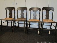 Front view of four dark painted wooden chairs with caned seats and gold stenciling on curved backrests, showing wear and some damage to seats