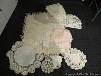 Assorted vintage linens and doilies including lace curtains, pink coasters, a tea cozy, various crochet and lace doilies in different shapes and sizes laid out on dark surface.