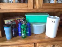 Photo of assorted miscellaneous plastic travel Tupperware items arranged on wooden cabinet shelf including water pitcher, thermoses, water bottles, rectangular and round containers.