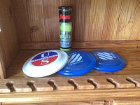 Photo showing tube of Penn tennis balls and three frisbees on wooden coat rack hung on wood panel wall