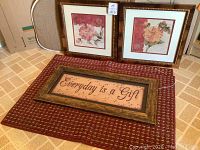 Three decorative framed pictures and two small rugs, displayed on the floor against a wall.