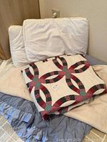 Two standard pillows and king size beige blanket folded and leaning against wall with a folded patchwork quilt placed on top