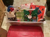 Red Rubbermaid storage tote filled with assorted Christmas gift bags, tissue paper, bows, ribbons, and boxes, showing variety of holiday wrapping supplies.